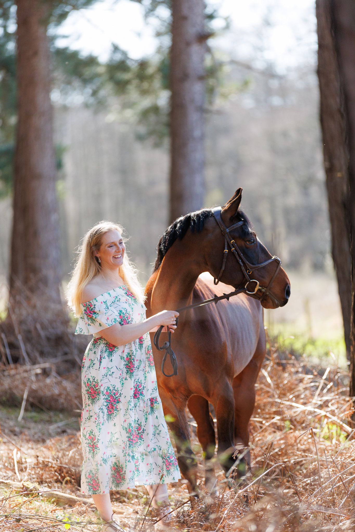 Smiling blonde woman in floral dress leading a bay horse through a sunlit forest.
