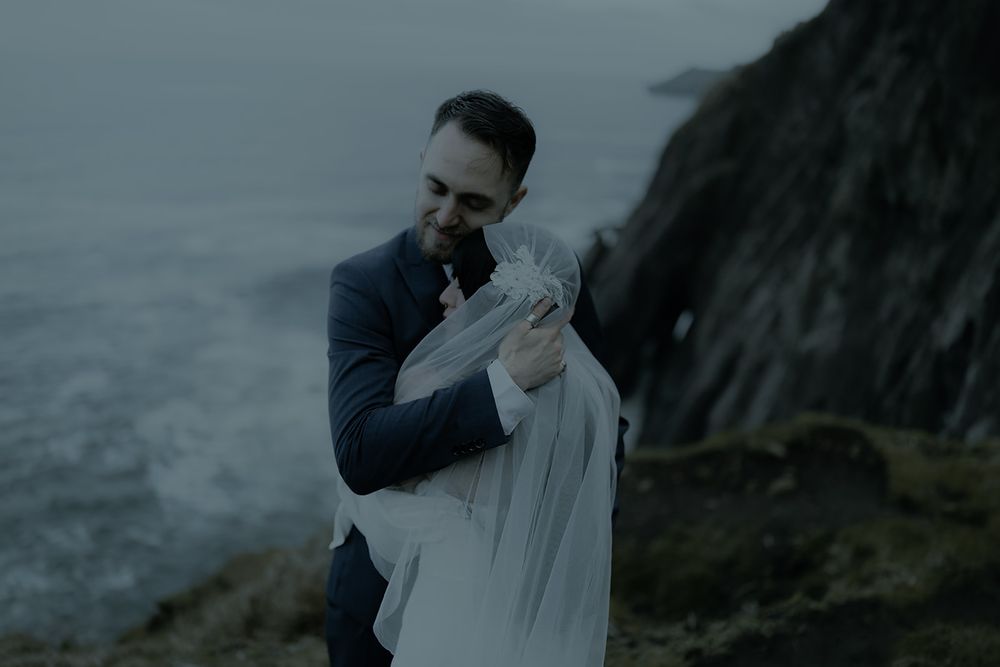 Couple embracing on a dramatic coastal cliff, groom in navy suit holding bride in flowing white veil, moody ocean backdrop.