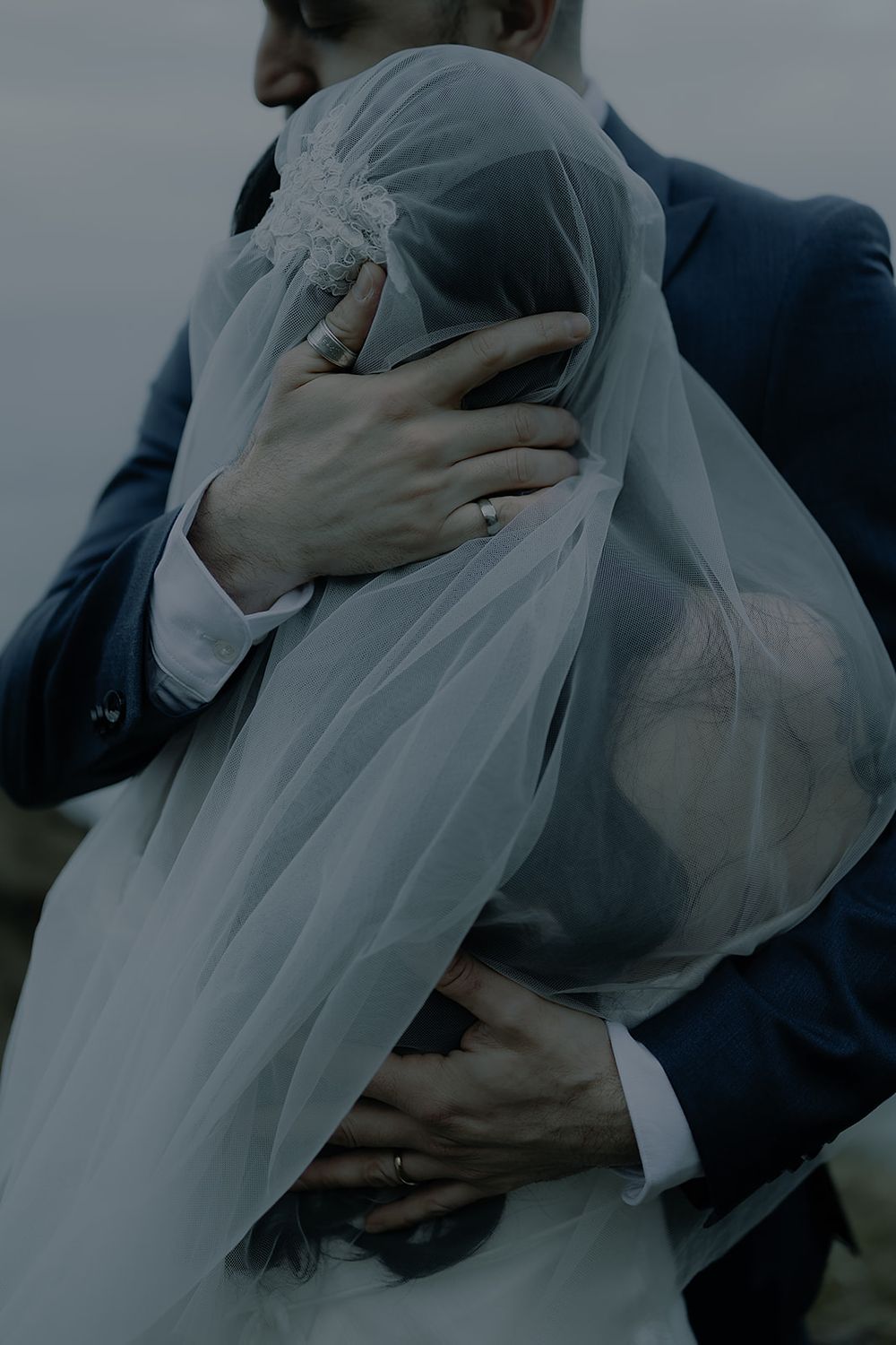 A groom in a navy suit embraces his bride, her face covered by a flowing white veil, both wearing wedding rings.