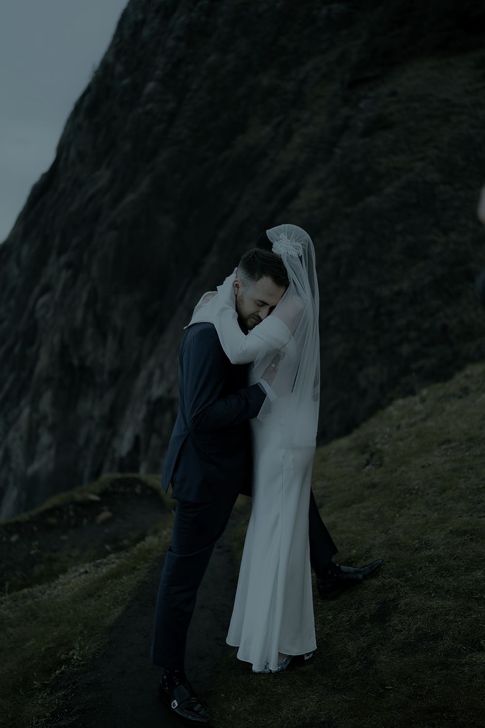 Bride and groom embracing on a dramatic misty hillside, bride in white gown and veil, groom in dark suit.