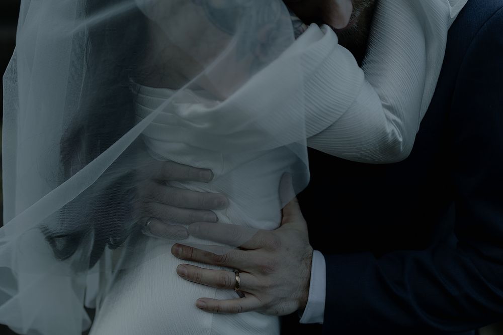 Close-up of groom's hand on bride's waist, bride wearing wedding ring, soft white veil draped over them.