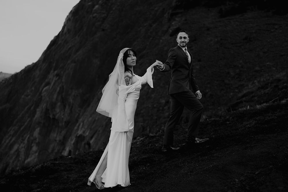 Black and white photo of a bride and groom holding hands on a rocky hillside during their outdoor wedding.