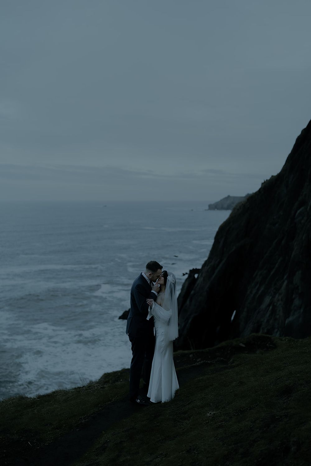 Bride and groom share a romantic moment on a dramatic coastal cliff at dusk, with moody ocean waves below.