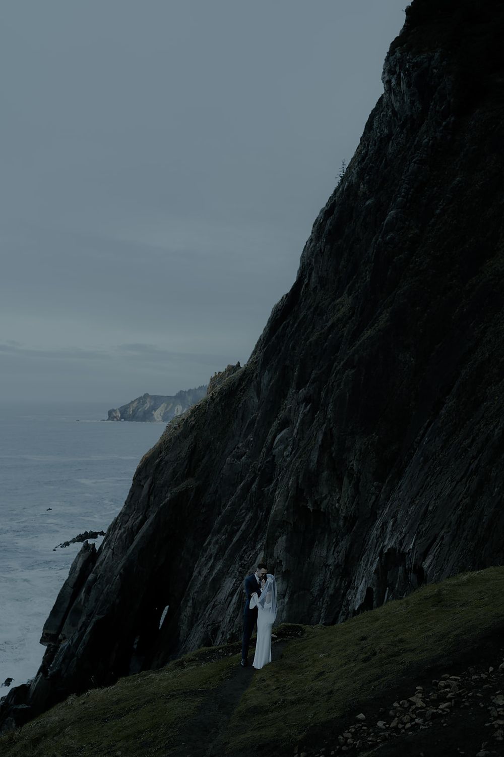 Couple in white wedding attire standing on dramatic coastal cliffs overlooking a misty ocean on an overcast day.
