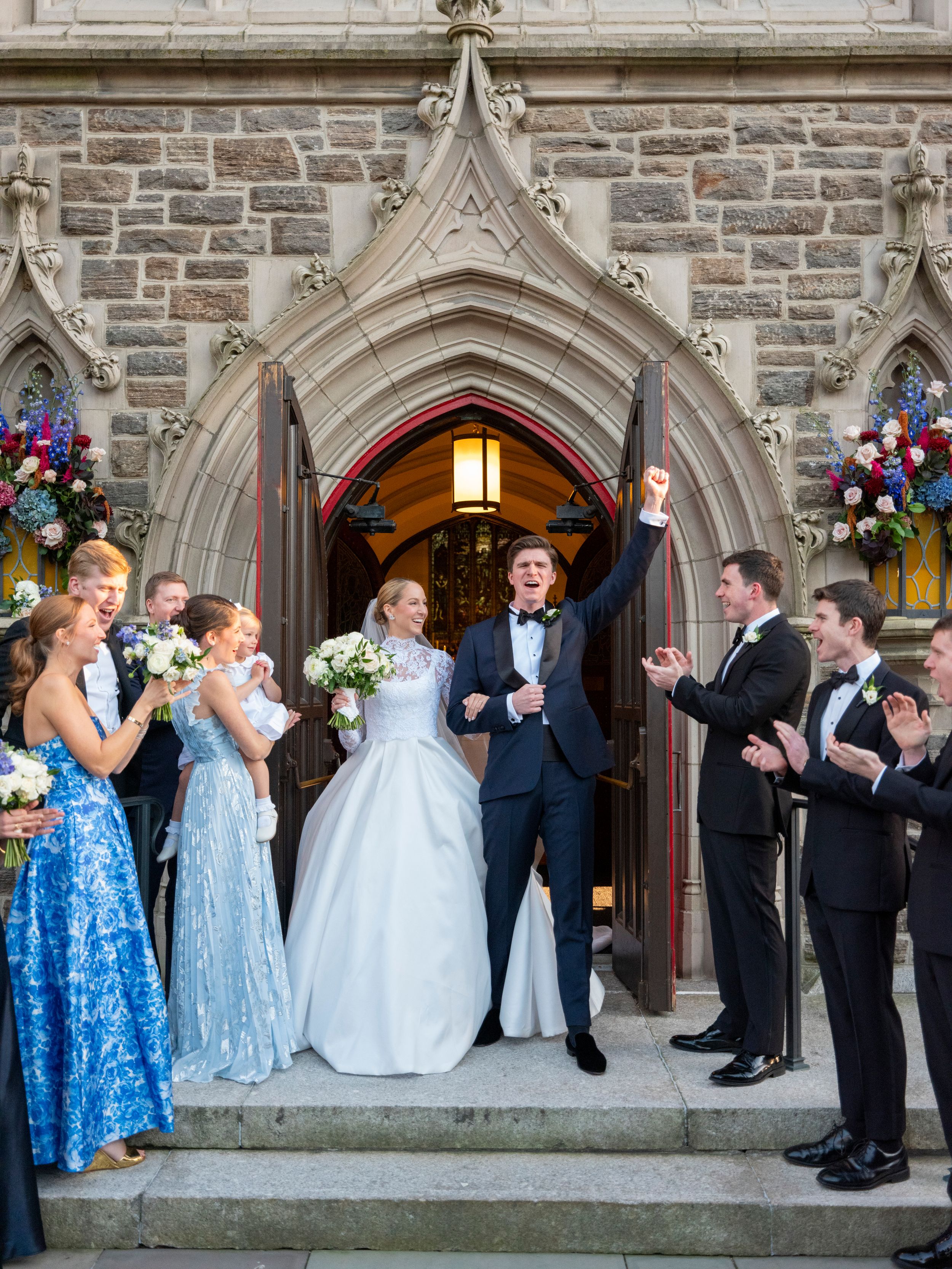 Wedding party in formal attire poses along stone church wall with Gothic arched doorways and stained glass windows.