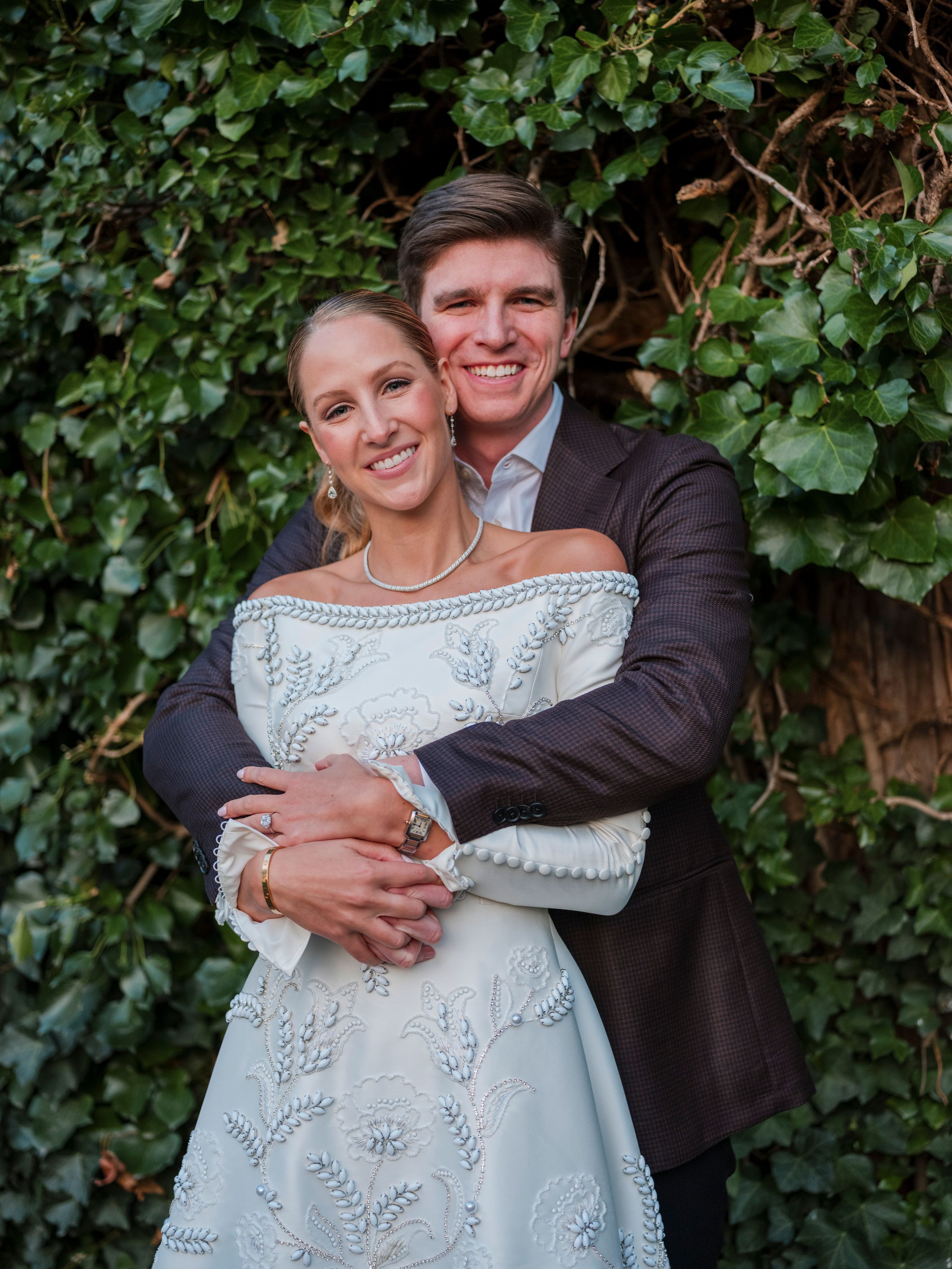 Couple embraces in front of lush green ivy wall wearing formal wedding attire with bride in white lace dress.
