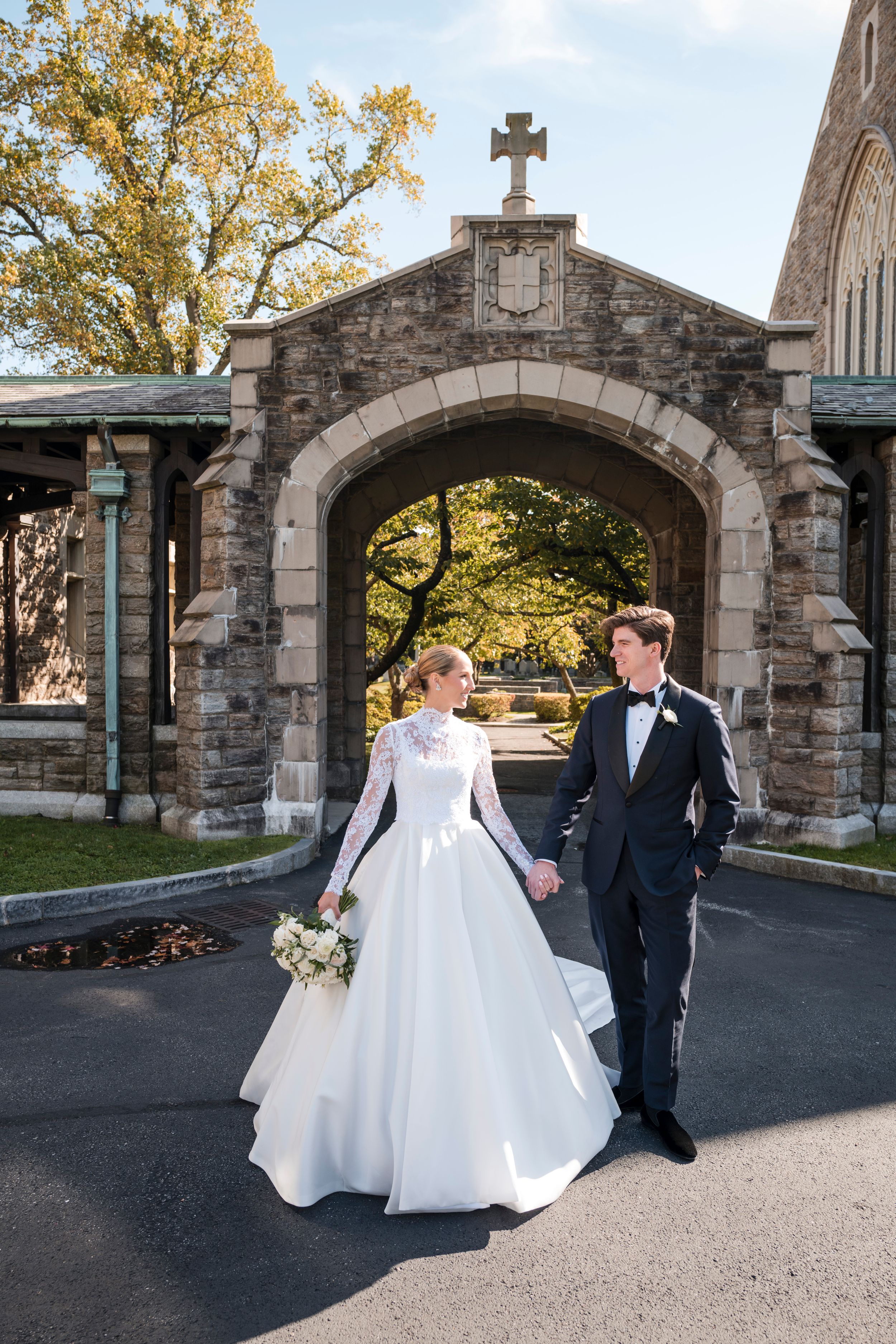 A couple in wedding attire strolls through a stone archway walkway during an autumn day.