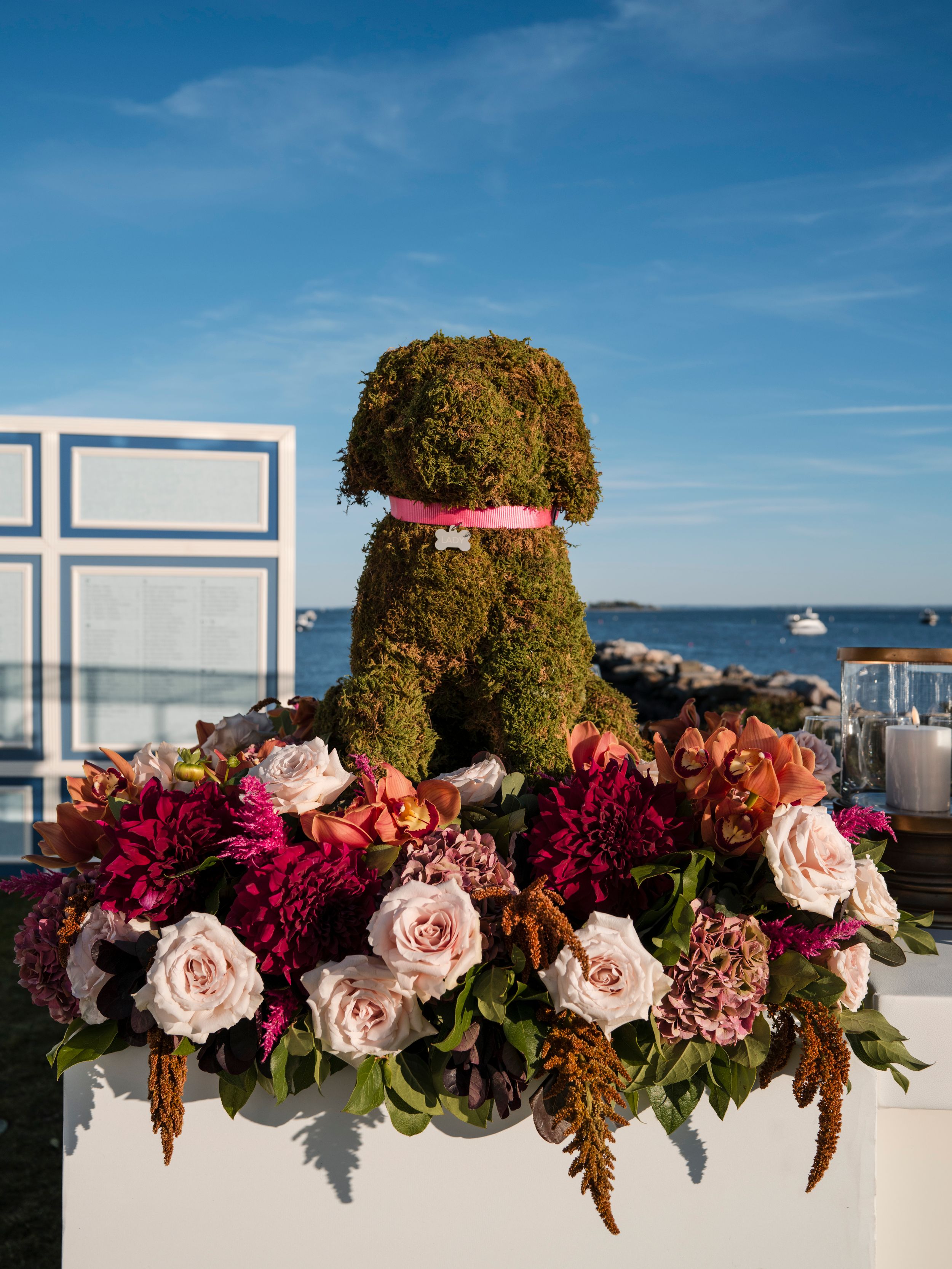 Elegant floral arrangement with pink roses, burgundy blooms, and dried foliage against a bright blue sky backdrop.
