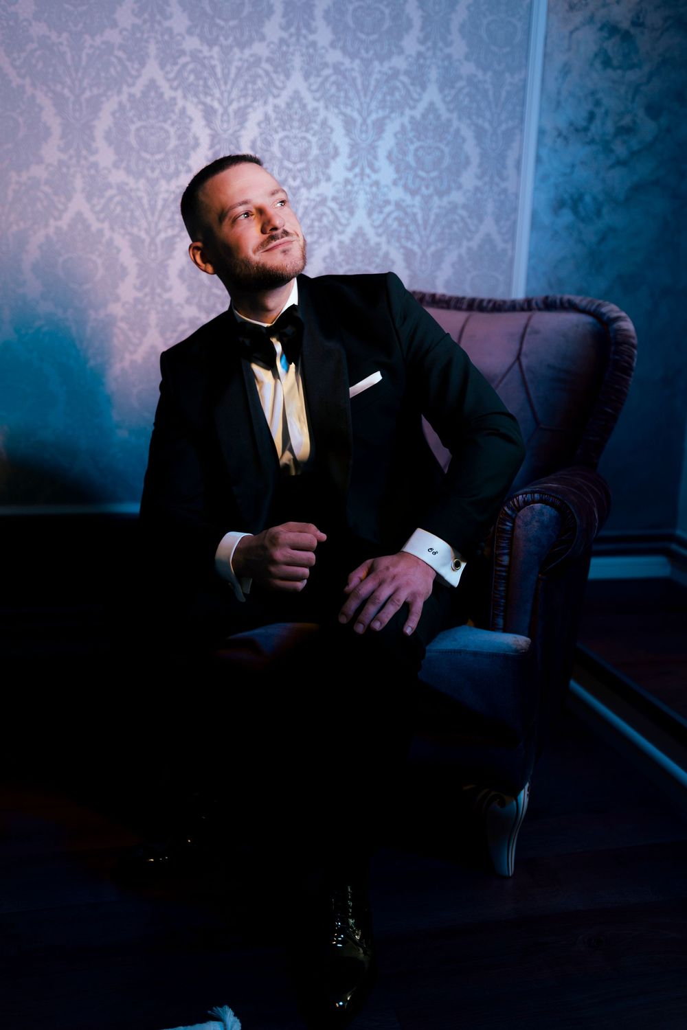 Elegant groom in black tuxedo with bow tie sits confidently in velvet armchair against ornate wallpaper.