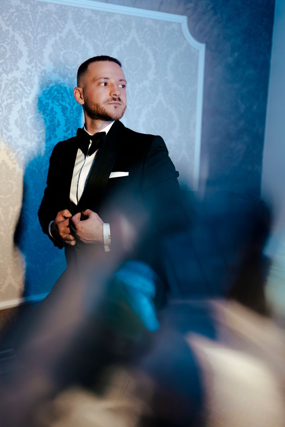 Stylish groom in black tuxedo stands against blue damask wallpaper in elegant wedding preparation room.