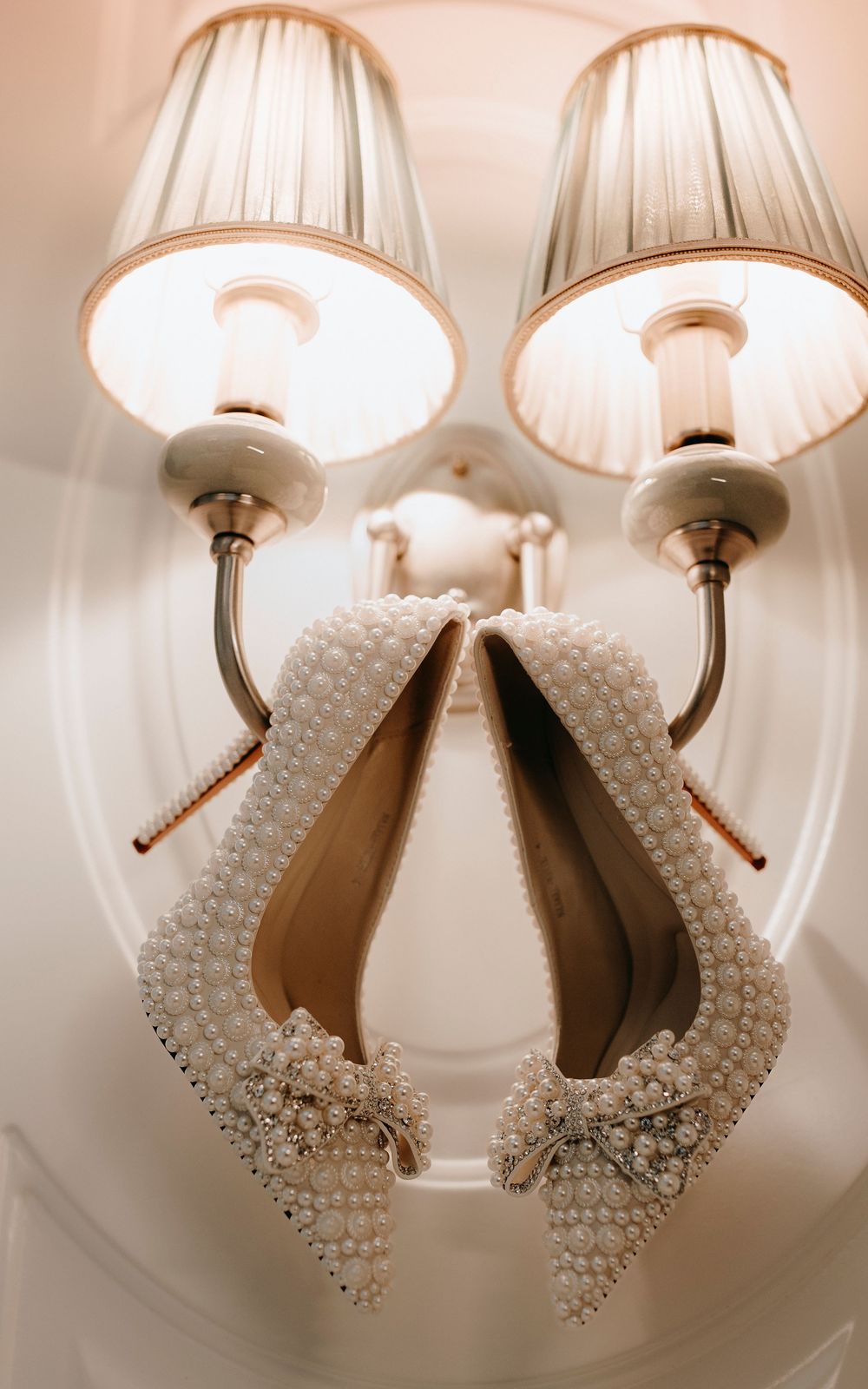 Pearl-encrusted bridal heels with bow detail hanging elegantly on a vintage wall sconce light fixture.