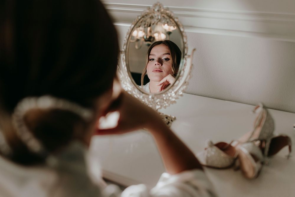 Bride in white robe applies makeup while gazing into ornate vintage mirror with bridal heels nearby.