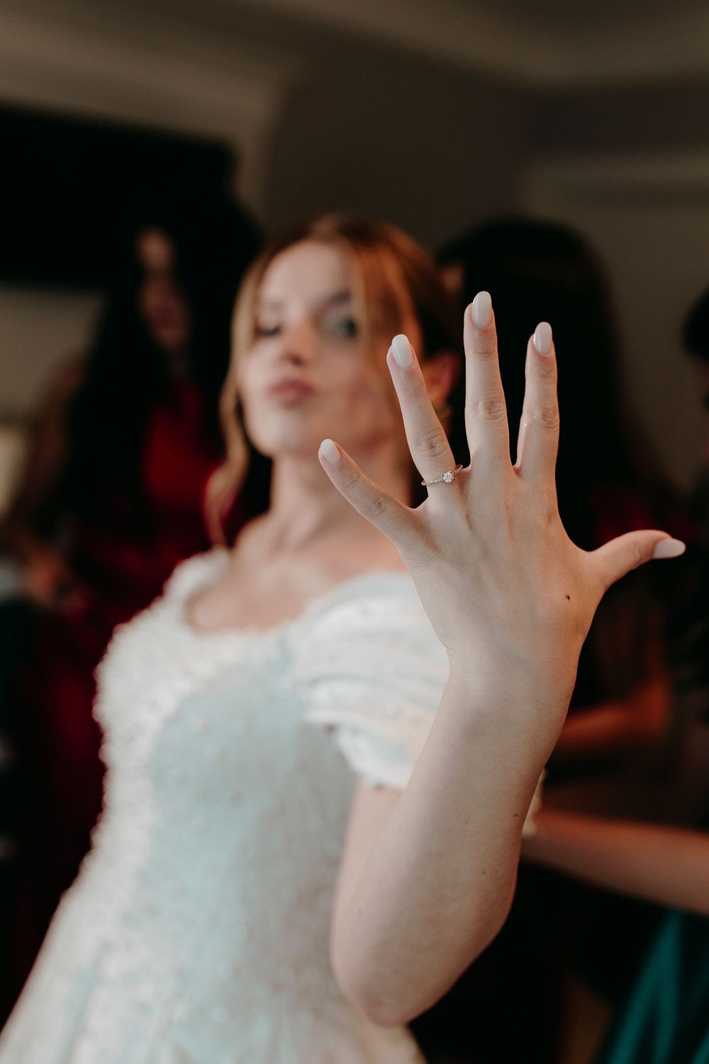 Bride in white gown showing off her diamond engagement ring to the camera with bridesmaids behind her.