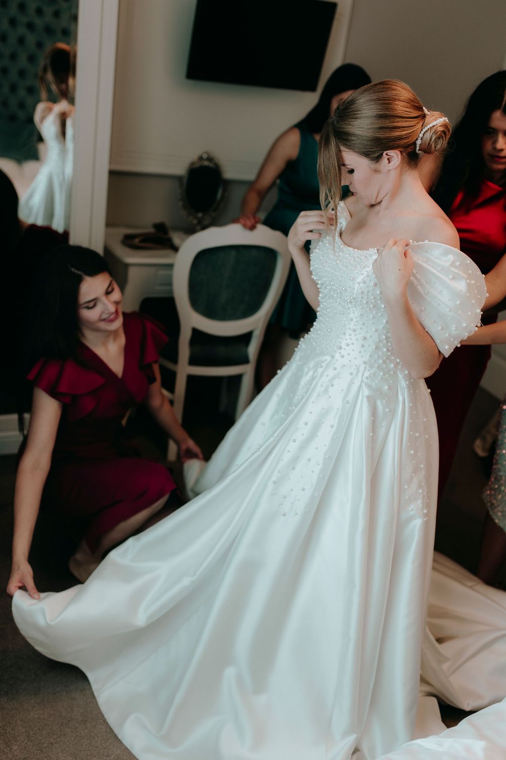 Bridesmaids in red dresses helping bride into her elegant off-shoulder pearl-embellished white wedding gown.