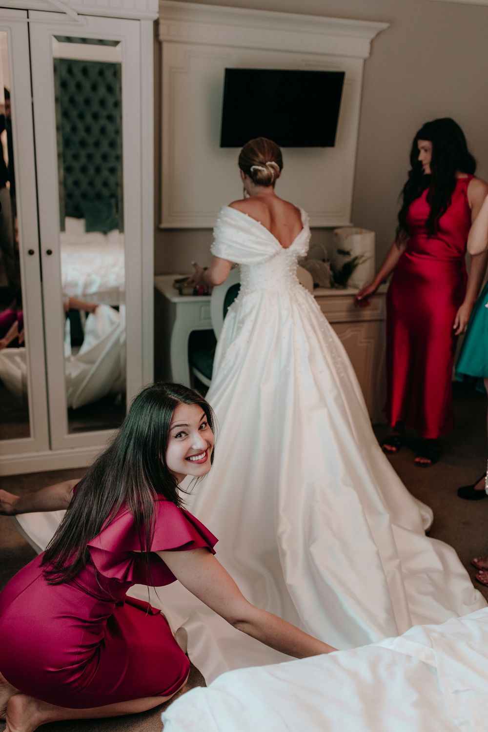 Bridesmaid smiling at camera while adjusting bride's white ball gown train in a bridal preparation room.