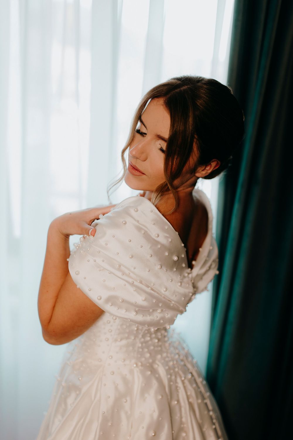 Bride in pearl-embellished white wedding gown adjusts dress near window with green curtains.