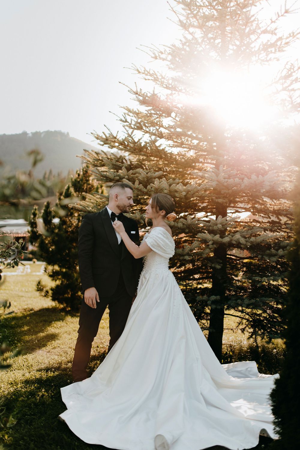 Bride and groom share a romantic moment outdoors near pine trees with golden sunlight glowing behind them.