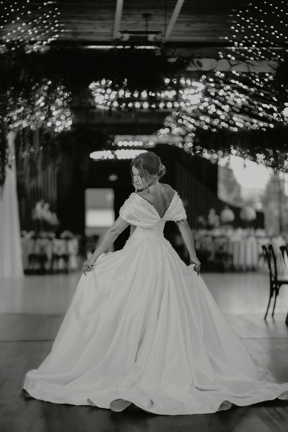 Black and white photo of a bride twirling in a ball gown inside a beautifully lit reception venue.