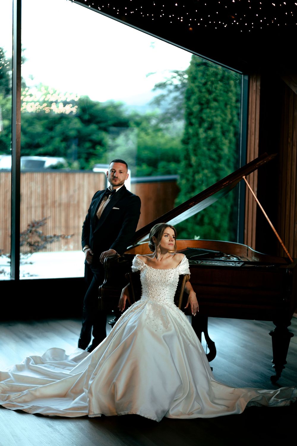 Elegant bride in a white ball gown seated by a grand piano with groom in black tuxedo standing behind her.