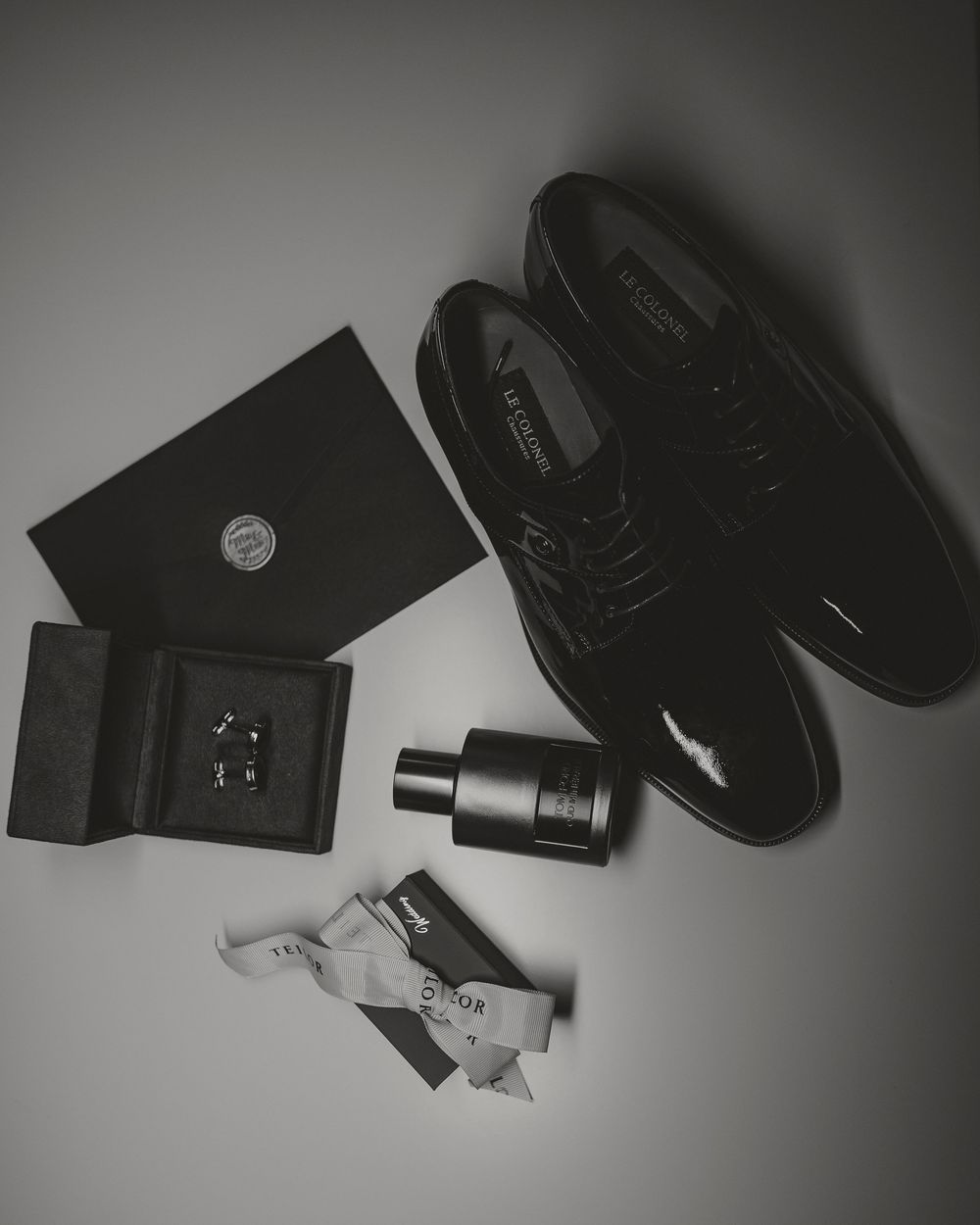 Black and white flat lay of groom accessories including dress shoes, cufflinks, and cologne.