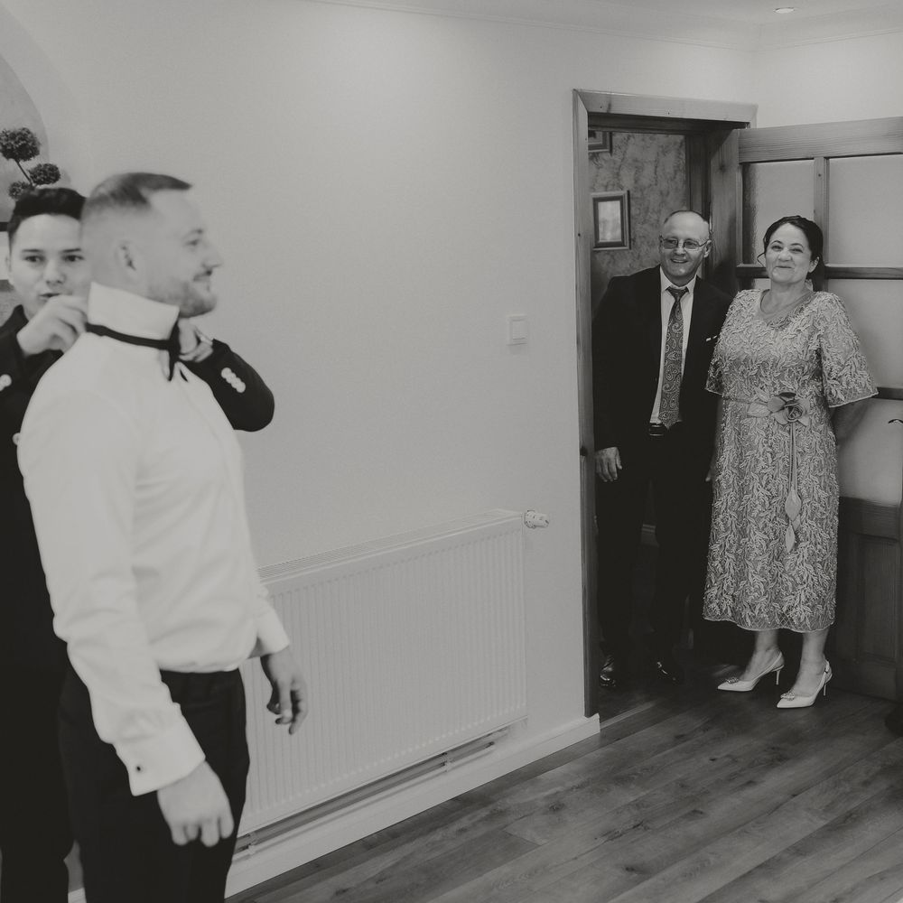 Black and white photo of wedding party getting ready as couple watches from doorway of elegant room.