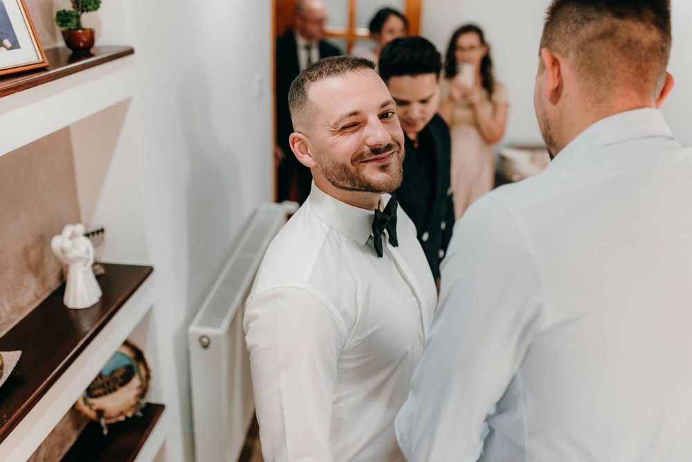 Smiling groom in white shirt with bow tie greeting guests before wedding ceremony indoors.