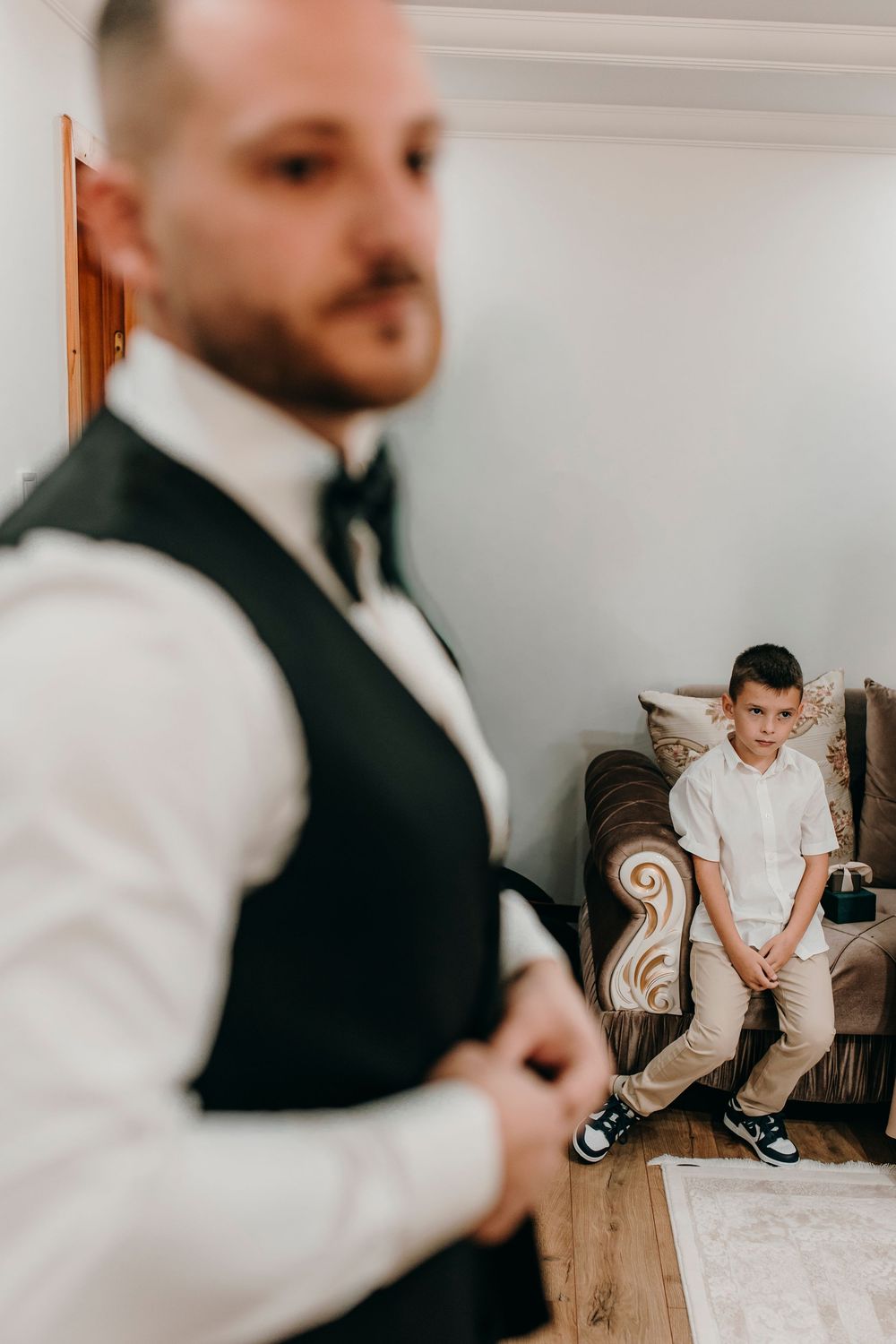 Groom in black vest and bow tie adjusts cufflinks while young boy in white shirt sits on sofa watching.