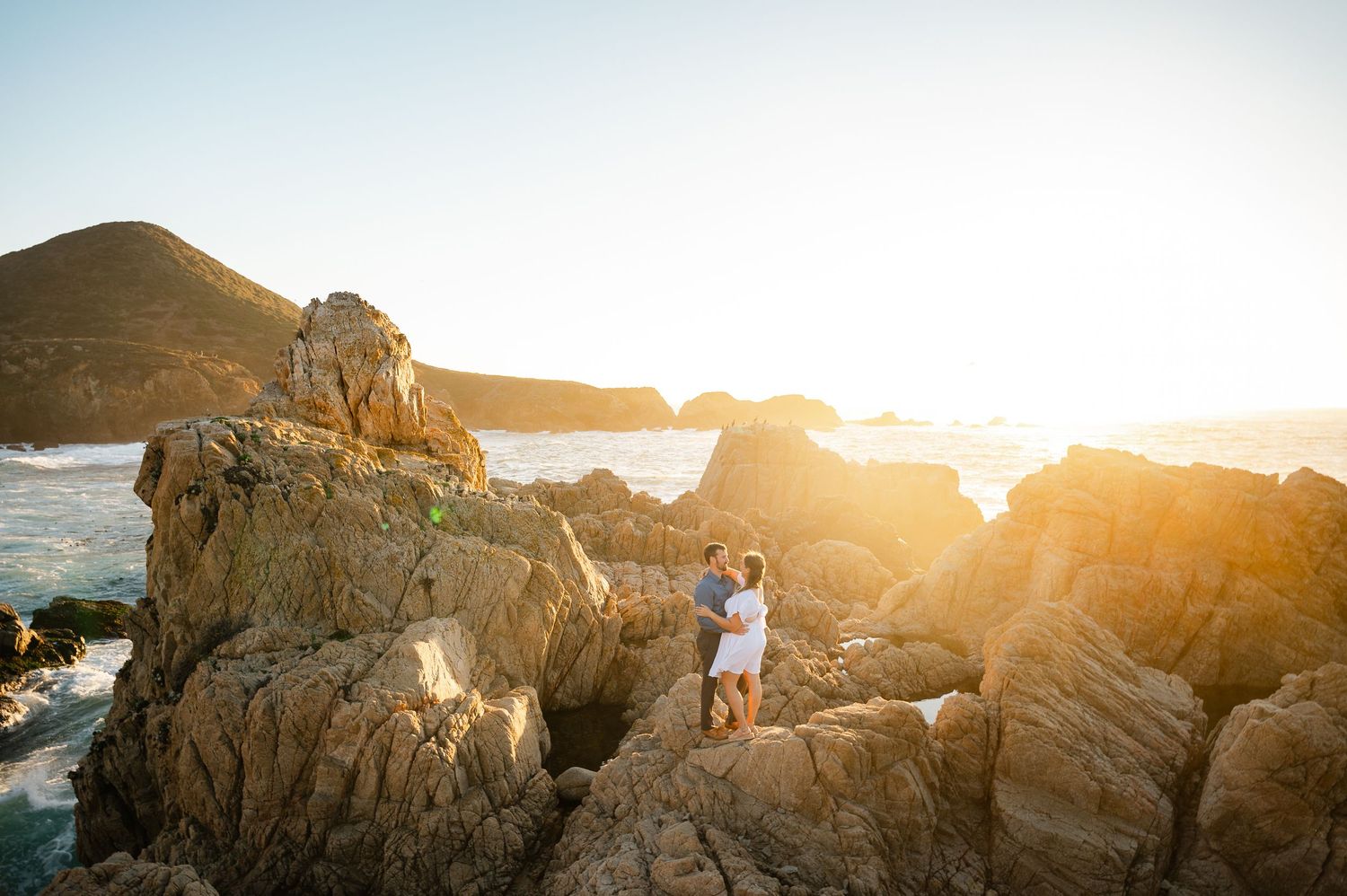 Couple embracing on golden sunlit rocky coastline at sunset with ocean waves and mountain backdrop.