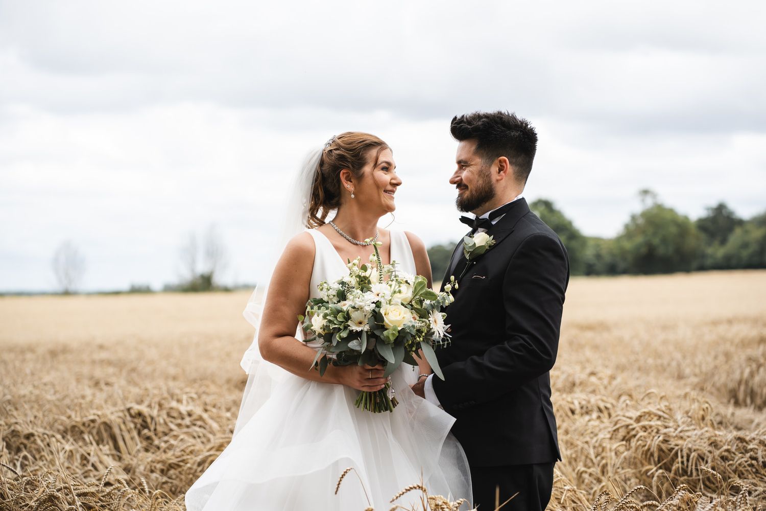 Laughing bride and groom holding a white bouquet standing together in a golden wheat field Lains Barn Wedding Venue