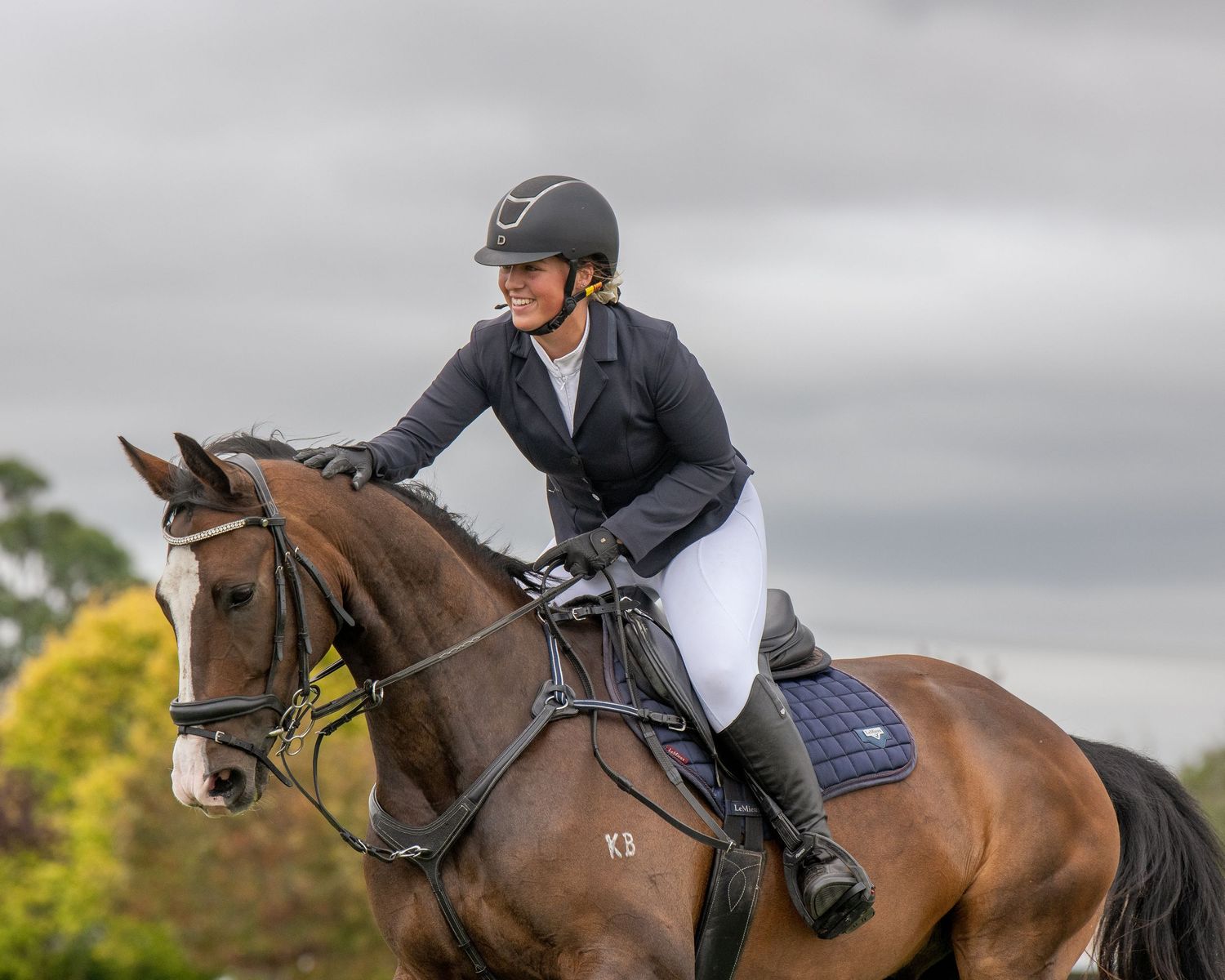 Female equestrian rider in black jacket and helmet competing on a bay horse at an outdoor show jumping event.
