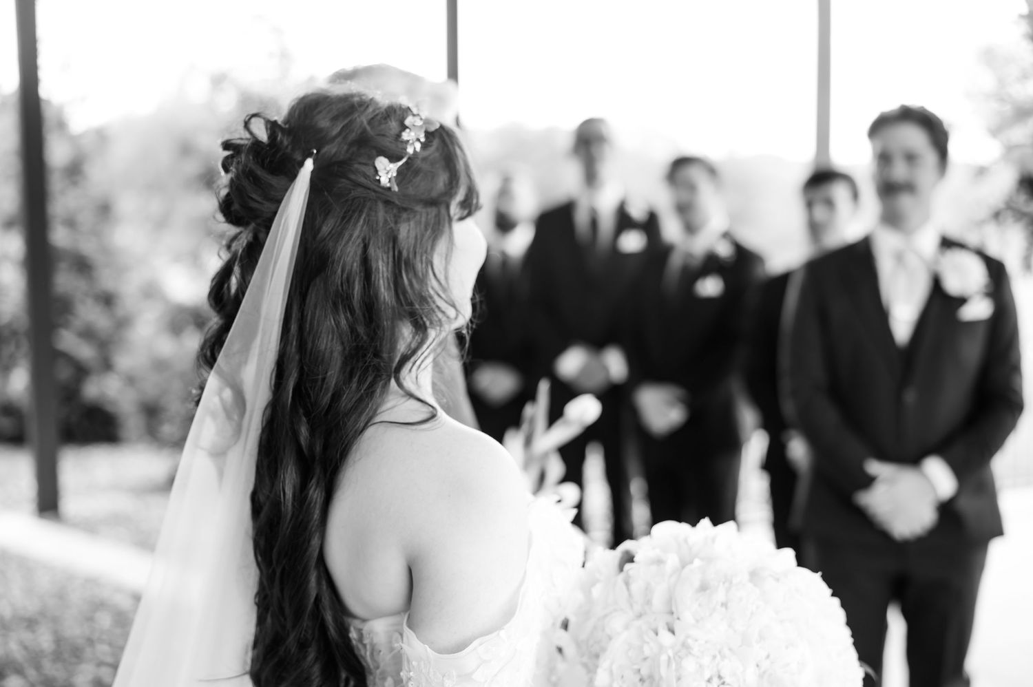 Bride holding bouquet faces groom and groomsmen in black suits at outdoor wedding ceremony, black and white photo.