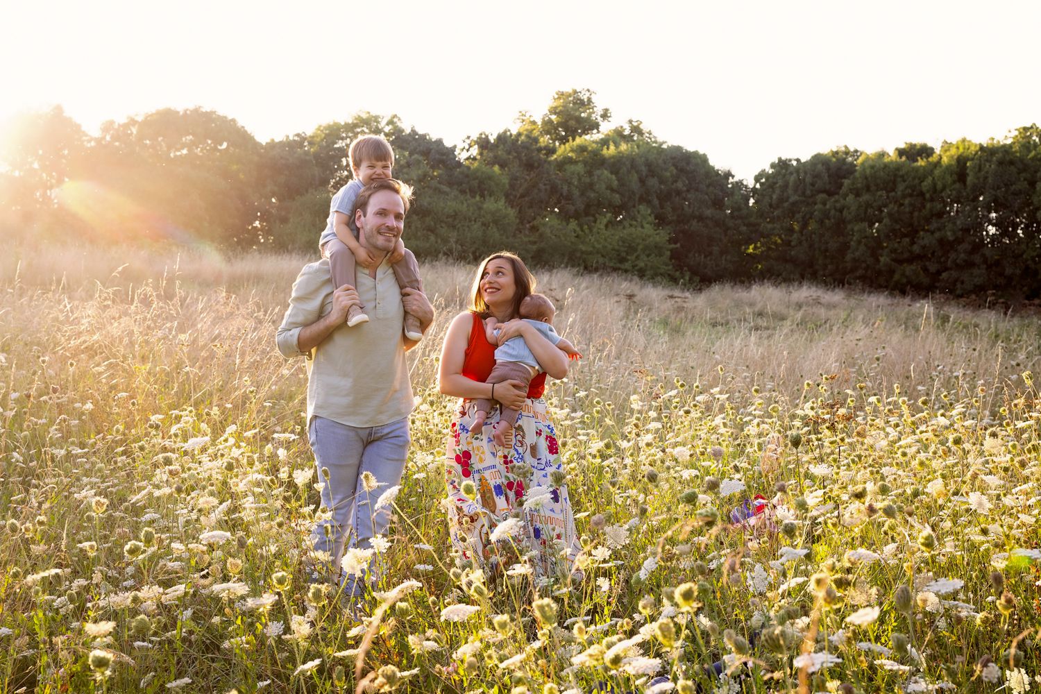 Family walking through sunny wildflower meadow, father carrying child on shoulders, mother holding baby, golden hour light.