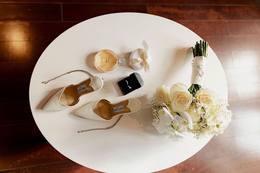 Wedding flat lay with bridal heels, ring box, bouquet of white roses, and accessories on white table.
