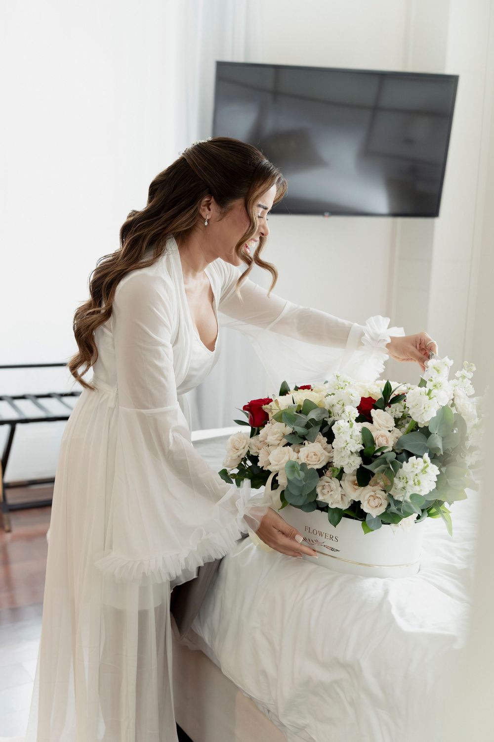 Smiling bride in white robe opening a floral gift box with red and white roses on her wedding morning.