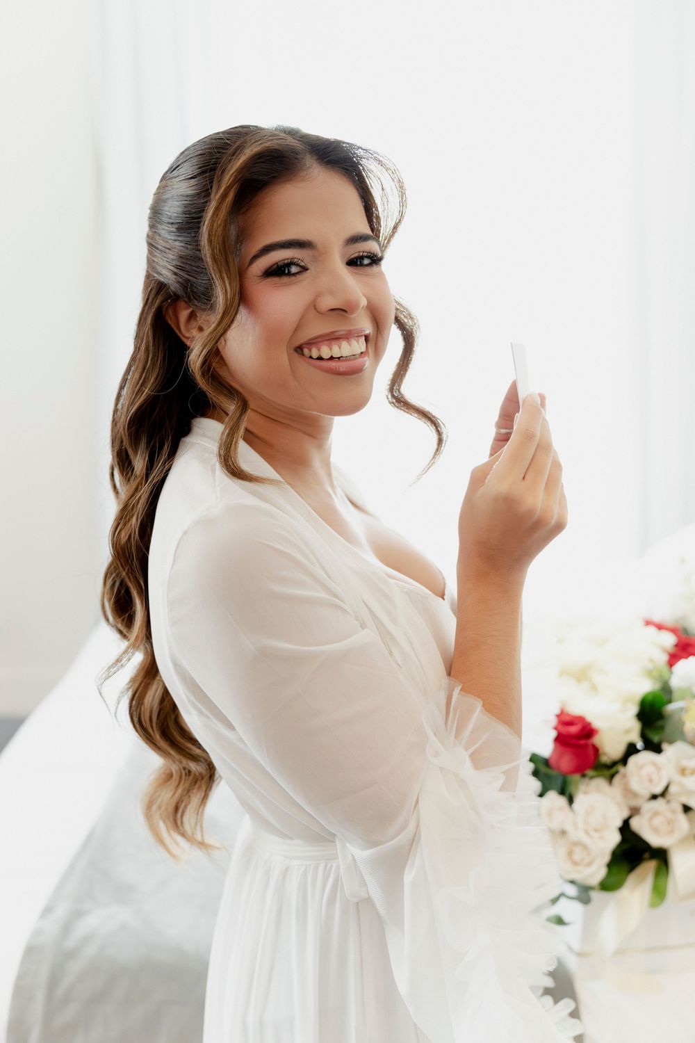 Smiling bride in white robe holding wedding card beside floral arrangement during bridal prep.