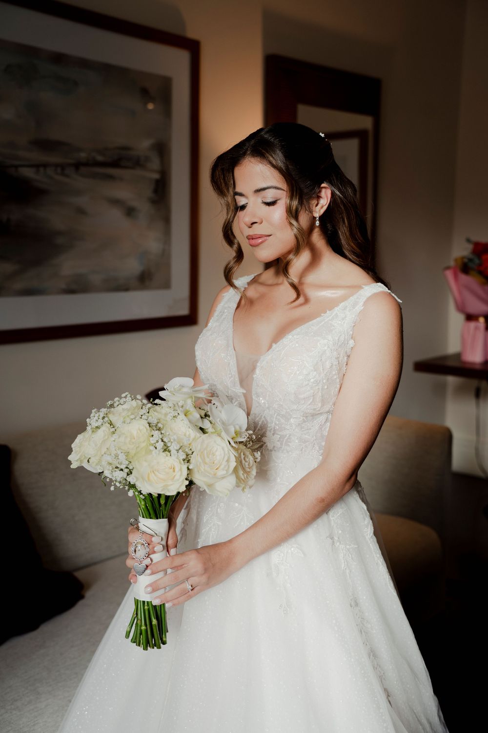 Elegant bride in lace ball gown holding white rose bouquet in softly lit hotel room.