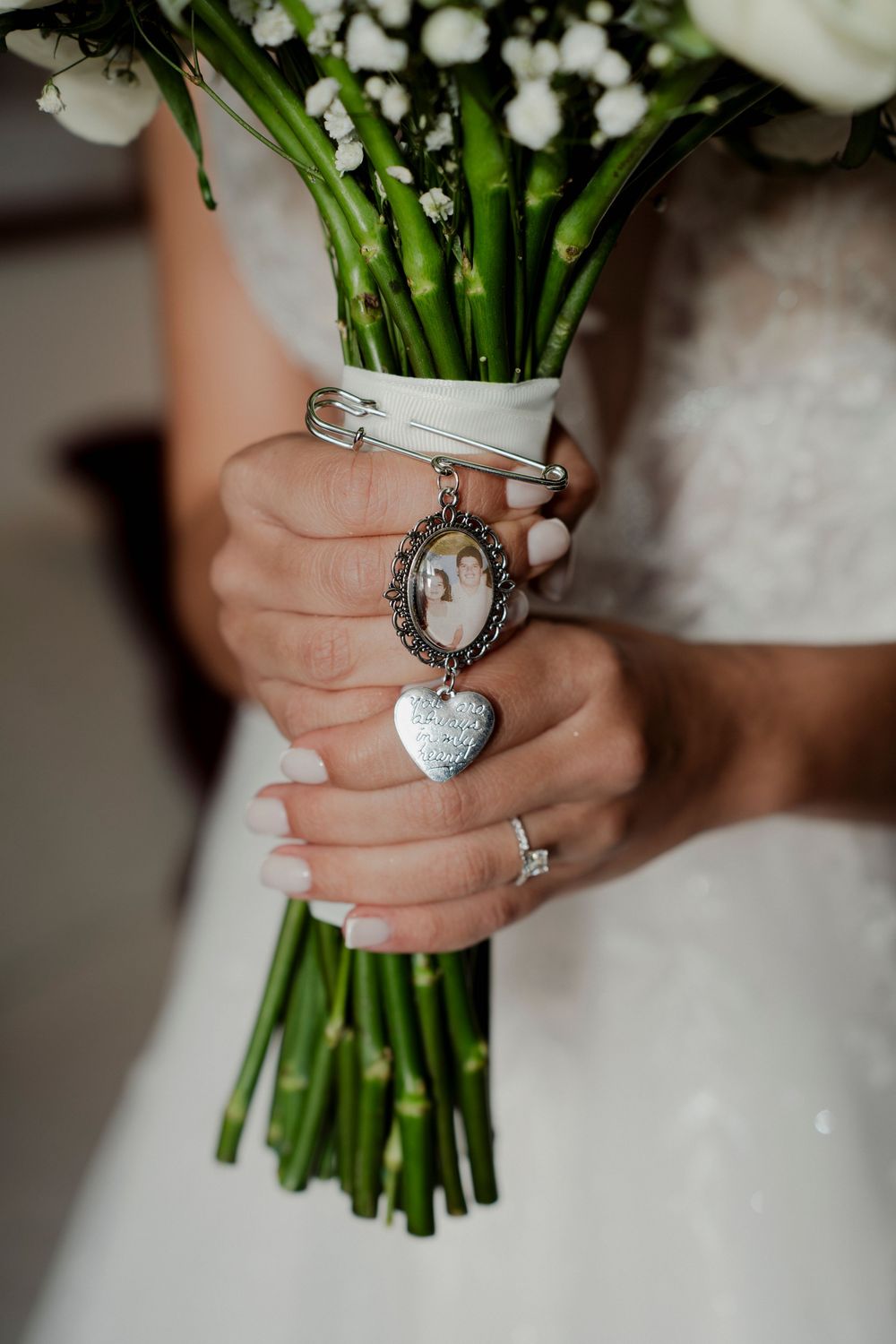 Bride holding white wedding bouquet with silver heart locket and photo charm memorial pin.