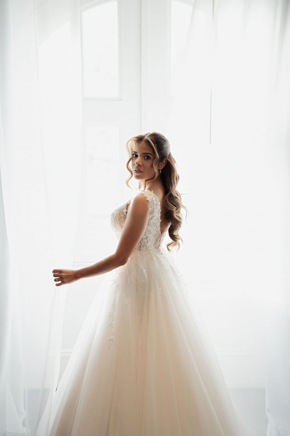 Elegant bride in a lace ballgown wedding dress standing by a bright white window, looking over shoulder.