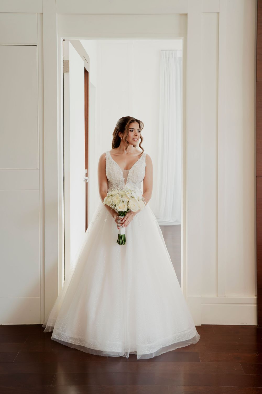 Bride in elegant white ball gown with lace bodice holding white bouquet standing in doorway.