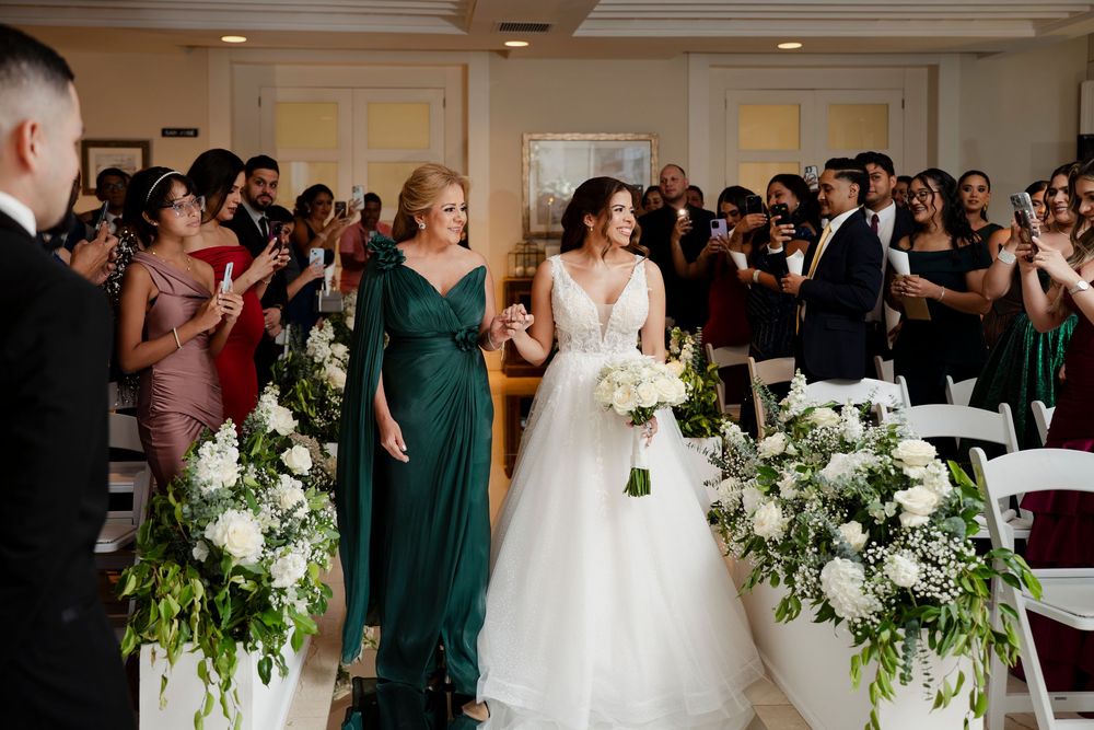 Bride walking down aisle in white gown escorted by woman in green dress at indoor ceremony.