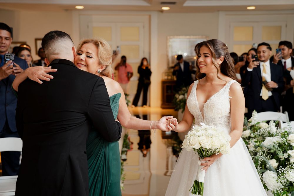 Bride smiling at wedding ceremony while guests celebrate in elegant white decorated venue.