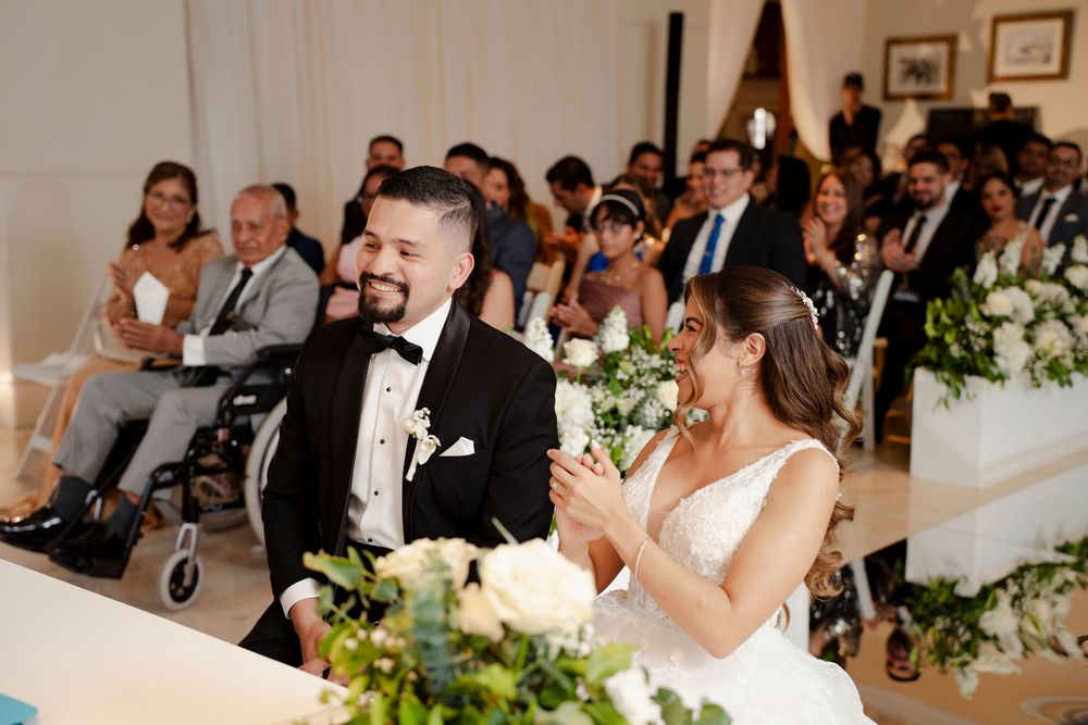 Smiling bride and groom at wedding ceremony altar with white floral arrangements and seated guests.