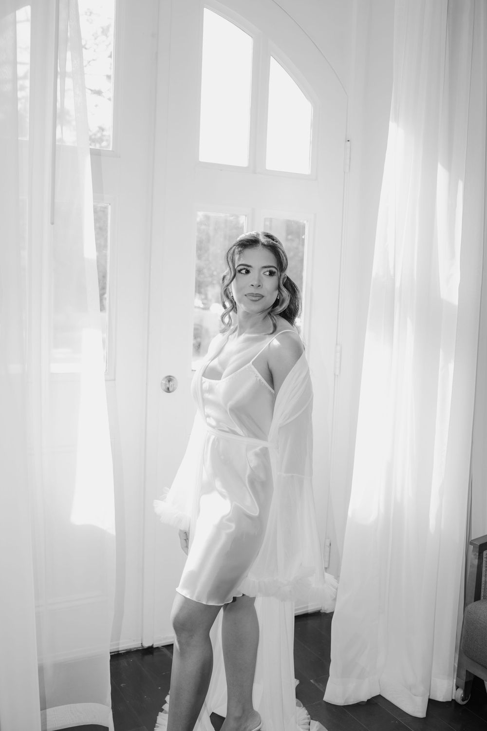Black and white photo of a bride in a short white robe standing in a doorway with sheer curtains.