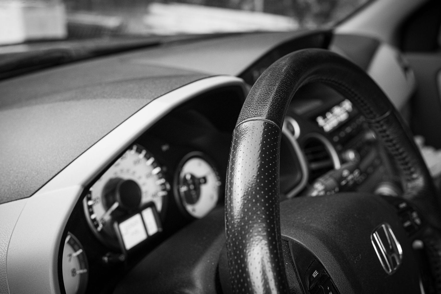 Black and white close-up of a car interior featuring a leather steering wheel and dashboard gauges.