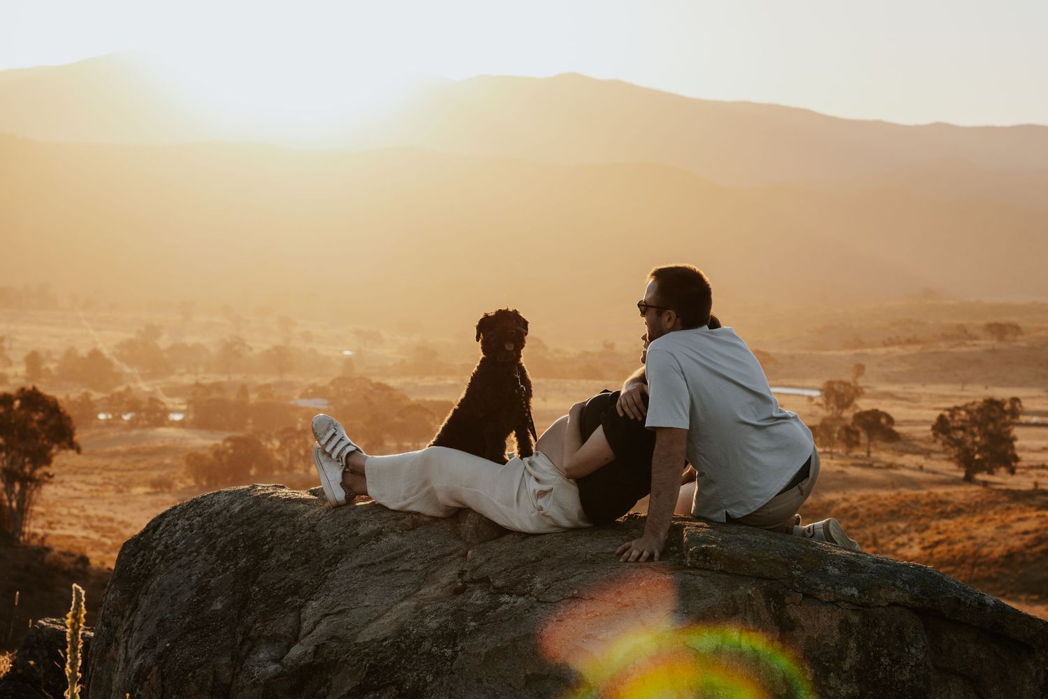 Couple and dog sitting on a rock watching a golden sunset over a scenic mountain valley landscape.