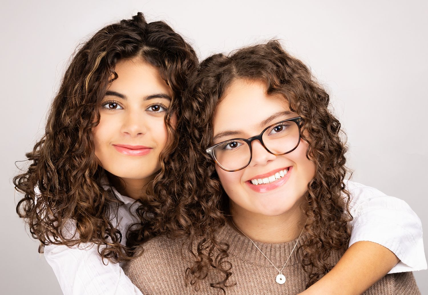 Two girls in front of a white/grey backdrop, smiling.  One has her arms wrapped around the other, in a landscape orientation