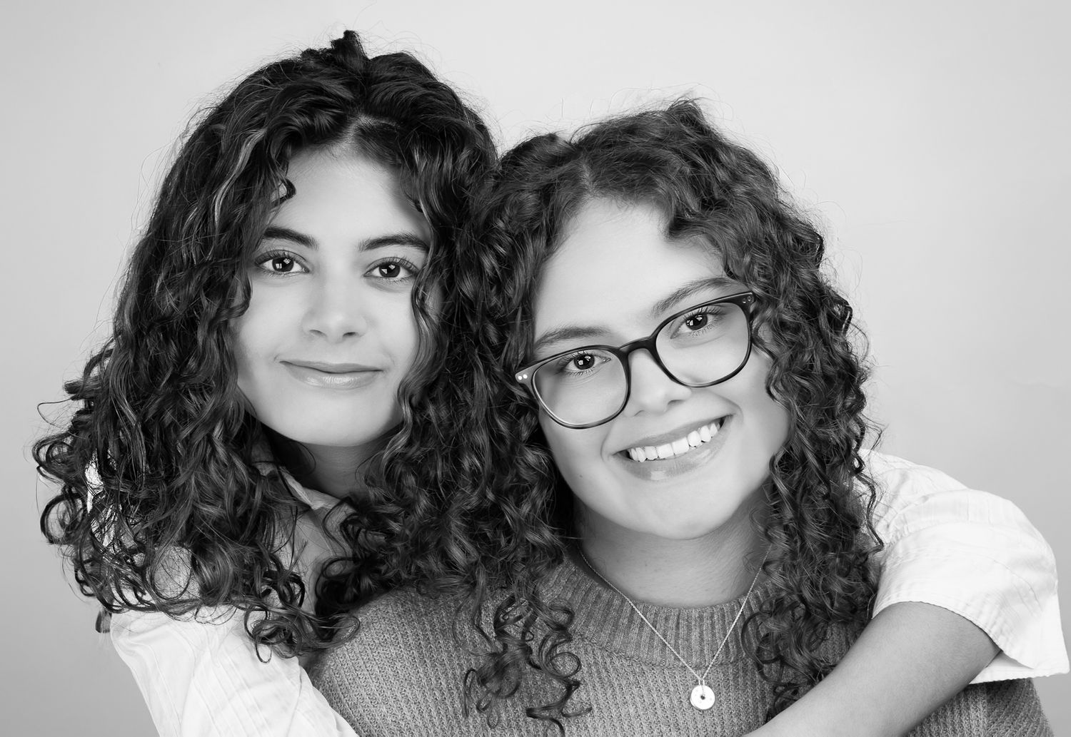 Black and white image of two girls in front of a white/grey backdrop, smiling.  One has her arms wrapped around the other