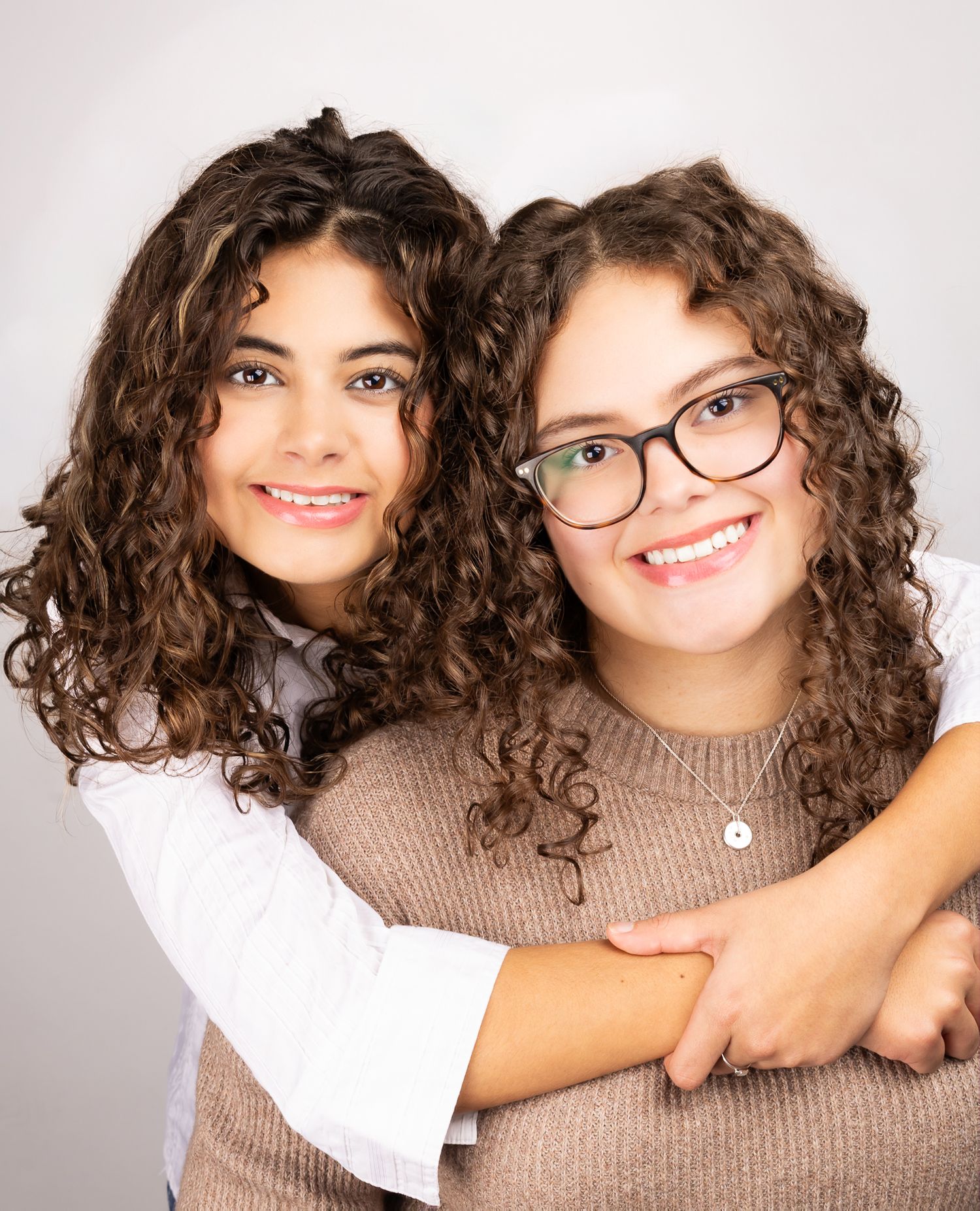 Two girls in front of a white/grey backdrop, smiling.  One has her arms wrapped around the other, portrait orientation