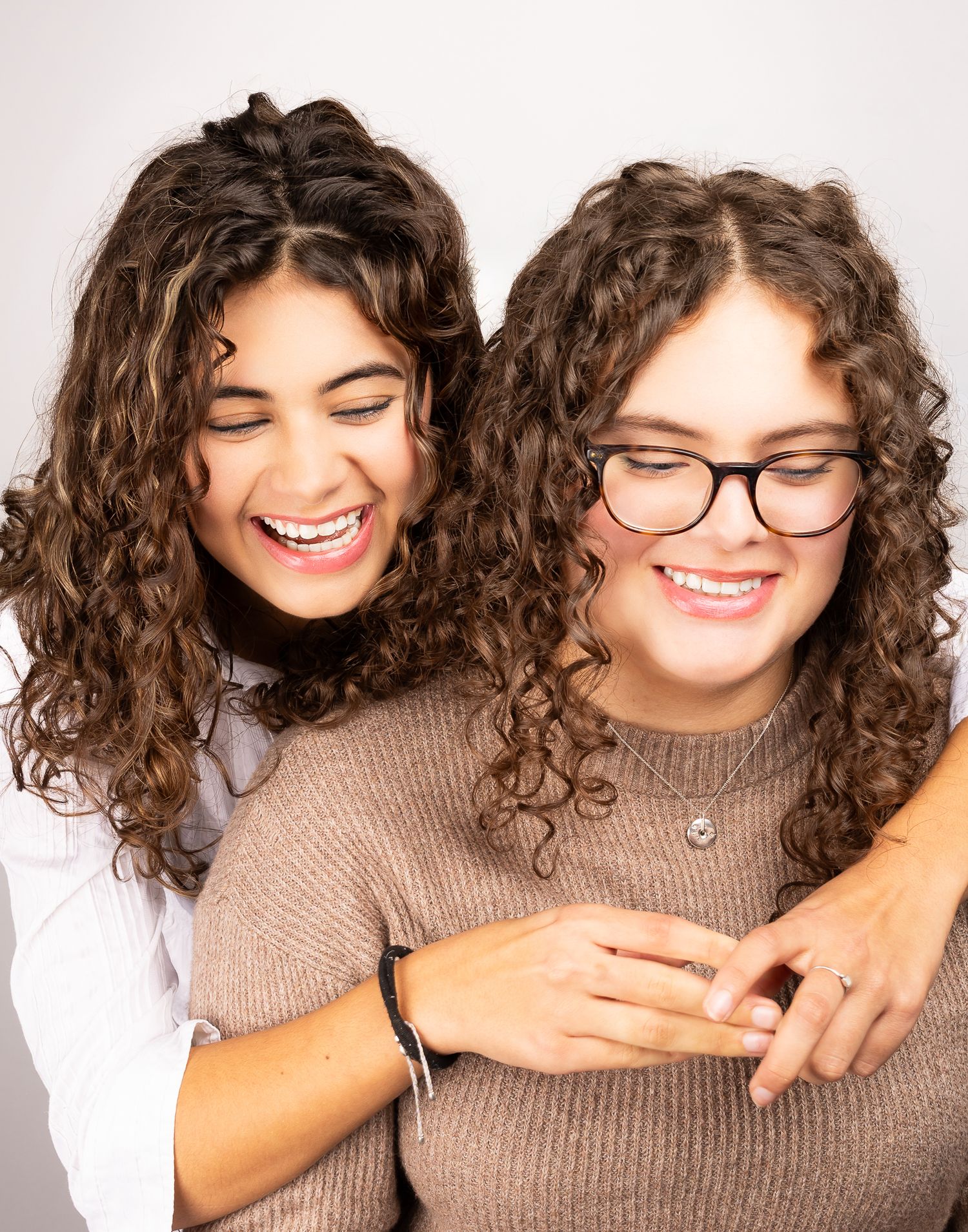 Two girls in front of a white/grey backdrop, mid-laugh.  One has her arms wrapped around the other, portrait orientation