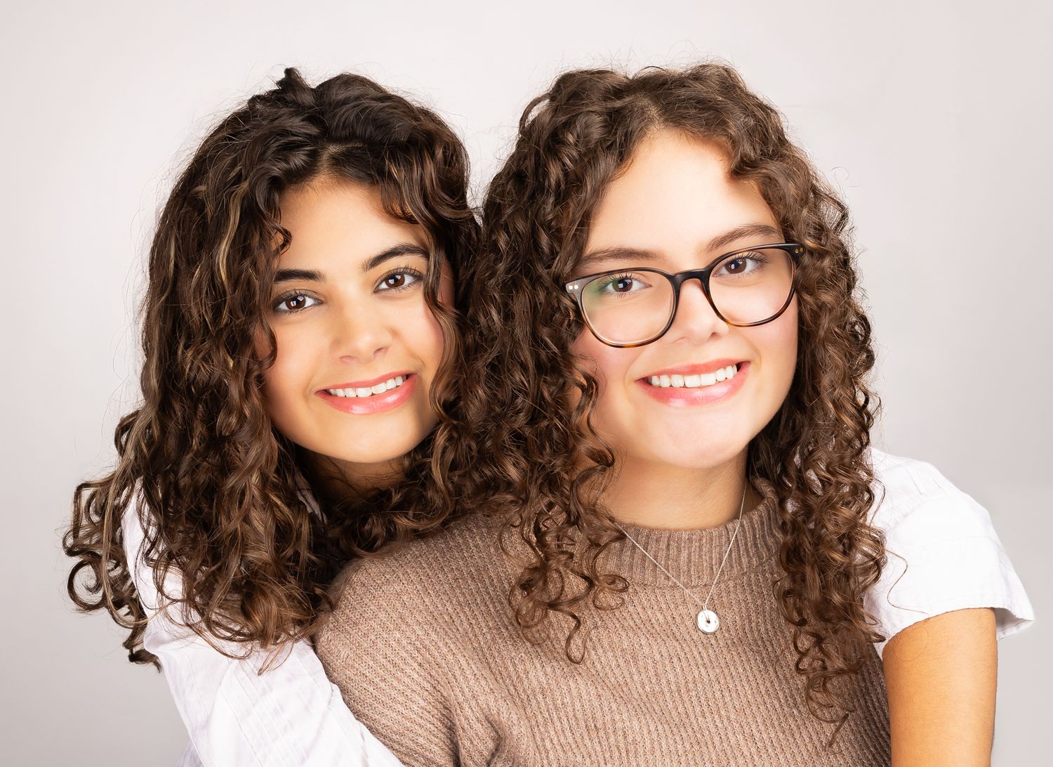 Image of two girls in front of a white/grey backdrop, smiling.  One has her arms wrapped around the other - quarter shot